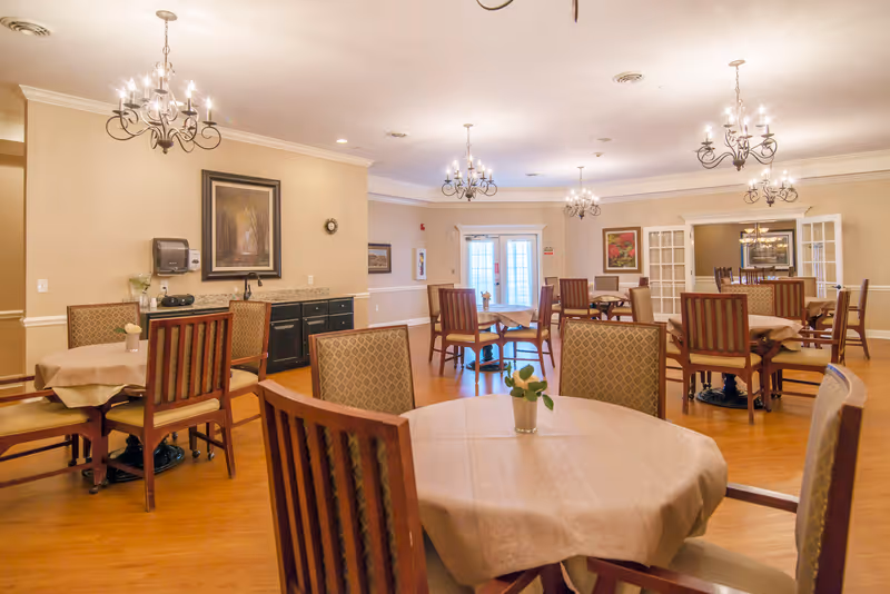 A spacious dining room with multiple round tables covered with beige tablecloths and surrounded by wooden chairs with cushioned seats and backs. The room features warm lighting from several ornate chandeliers hanging from the ceiling. The walls are painted a soft beige and decorated with framed artwork. There is a black sideboard against one wall with a paper towel dispenser and a small stereo. The floor is wooden, and French doors are visible in the background.