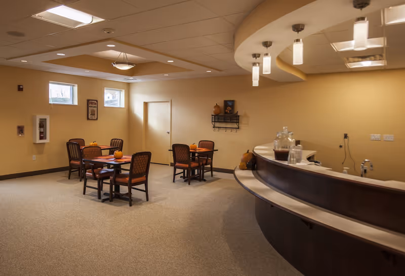 Communal dining area with small tables and chairs, pumpkins on the tables, and a curved service counter in a senior living facility.