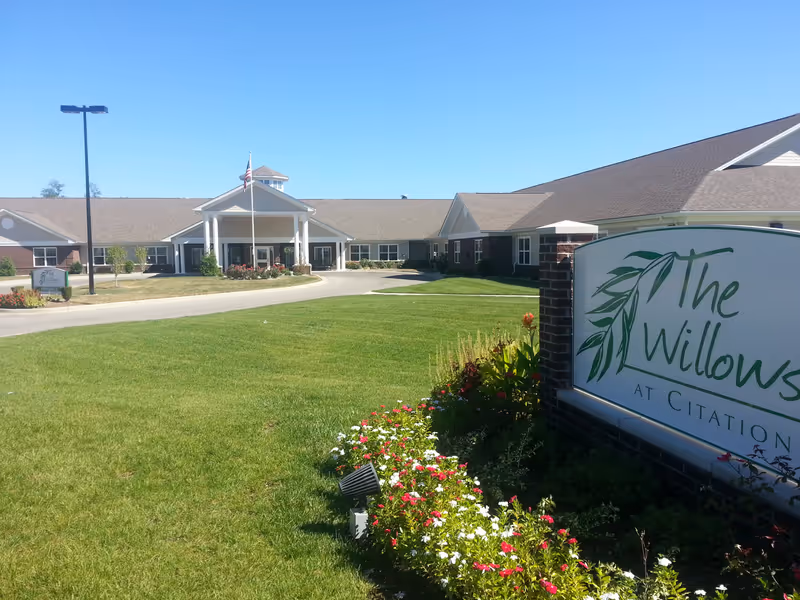 Exterior view of The Willows at Citation senior living facility with a large green lawn, flower beds, and a clear blue sky. The building has a covered entrance with white pillars and an American flag flying on a flagpole.