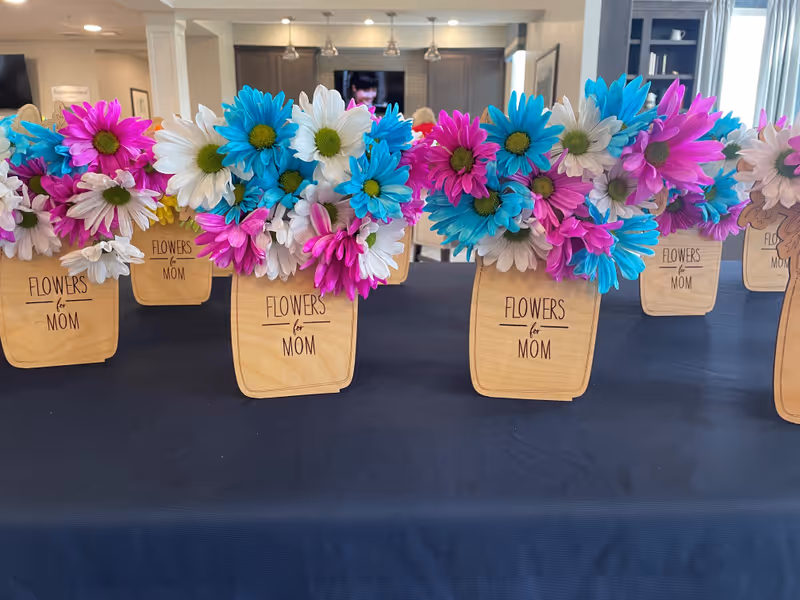 Several small wooden vases filled with colorful artificial flowers in pink, blue, and white arranged on a black tablecloth. Each vase has the words 'FLOWERS for MOM' written on it. The background shows a bright interior space with kitchen cabinets and a television.