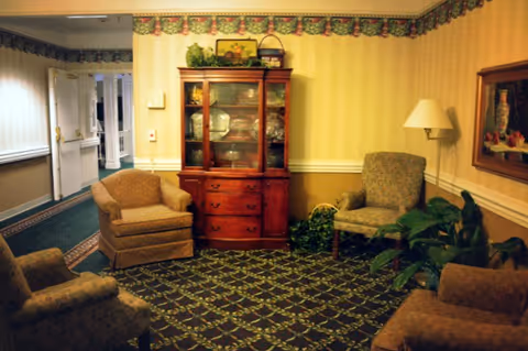 A cozy living room area in an assisted living facility with four upholstered armchairs arranged around a patterned carpet. There is a wooden china cabinet with glass doors displaying dishes, a floor lamp, a framed painting on the wall, and some potted plants. The walls have striped wallpaper with a floral border near the ceiling.