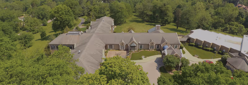 Aerial view of Cherokee Park Rehabilitation facility surrounded by lush green trees and landscaped grounds, showing multiple connected buildings with pitched roofs and a circular driveway at the entrance.