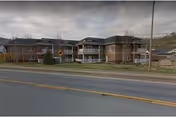 Front exterior of a two-story brick residential building with balconies, a lawn, and a road in front under a cloudy sky.
