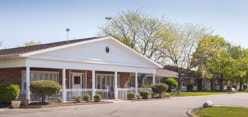 Front exterior of a single-story brick senior living building with a covered porch, columns, shrubs, and trees.