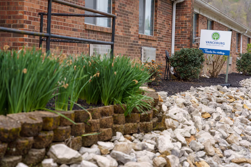Outdoor view of a brick building with a landscaped garden featuring green plants and rocks. A sign near the building reads 'Vanceburg Rehabilitation and Care, Thanks for visiting.'