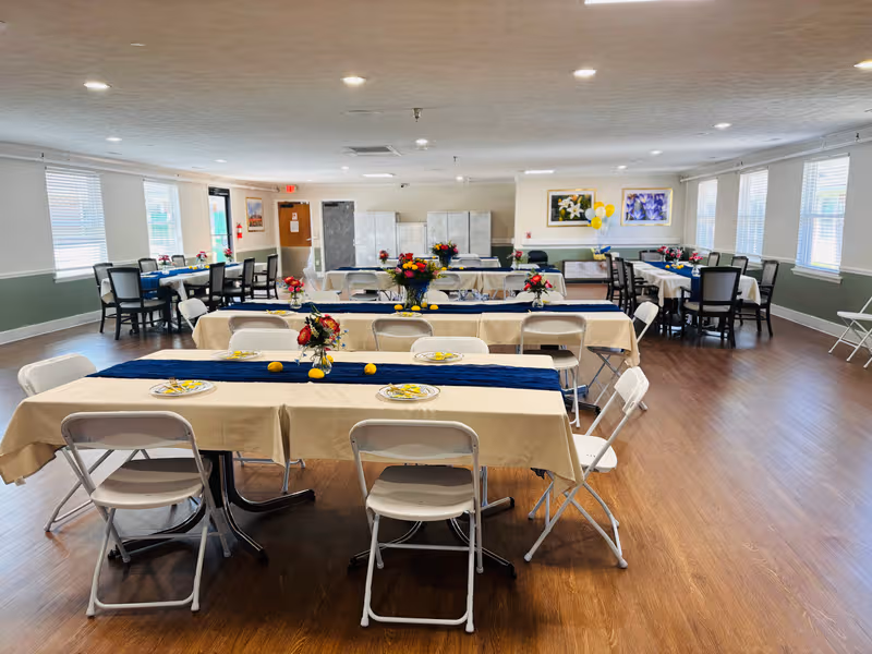 A spacious dining hall with long tables, beige tablecloths, navy runners, and floral centerpieces.