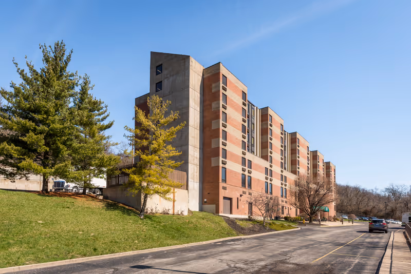 Exterior view of a multi-story brick and concrete senior living facility building with several windows, surrounded by green grass, trees, and a paved driveway under a clear blue sky.