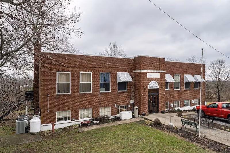 Two-story brick building front with a sign reading 'Jonesville Rest Home' above the entrance, window awnings, a small lawn and a red pickup truck parked nearby.