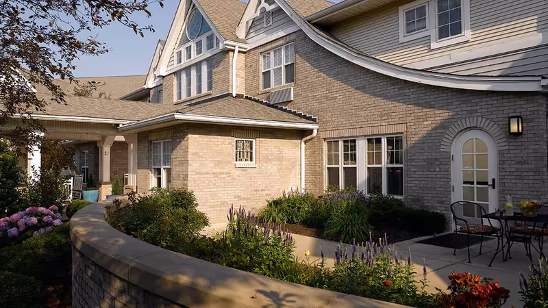 Exterior view of a senior living facility with beige brick walls, multiple windows, a curved stone wall surrounding a garden with various plants and flowers, and a small patio area with a table and chairs.