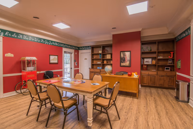 A bright communal activity room with a wooden table surrounded by chairs, bookshelves, a desk, and a popcorn machine against red walls.