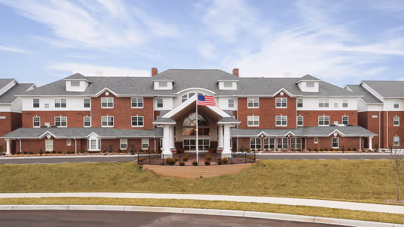 Front exterior of a three-story red-brick retirement community with a covered porte-cochère entrance, American flag, and landscaped lawn.