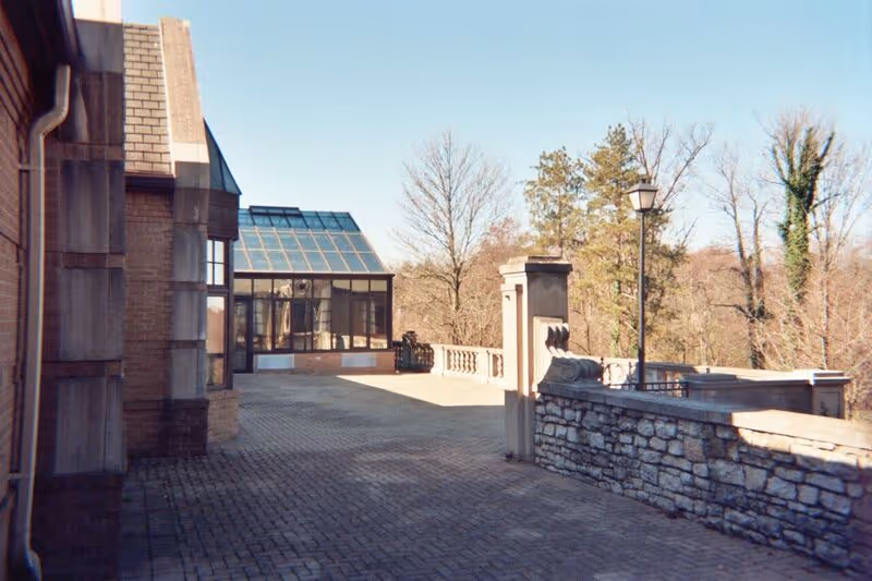 Outdoor paved terrace area adjacent to a brick building with a glass-enclosed sunroom or conservatory. Stone railing and lamp post line the edge of the terrace, with trees and clear sky in the background.
