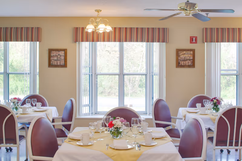 A bright dining room with round tables covered in white and yellow tablecloths, set with plates, cups, glasses, and silverware. Each table has a small vase with colorful flowers. The room has large windows with striped valances, framed pictures on the walls, and a ceiling fan with lights.