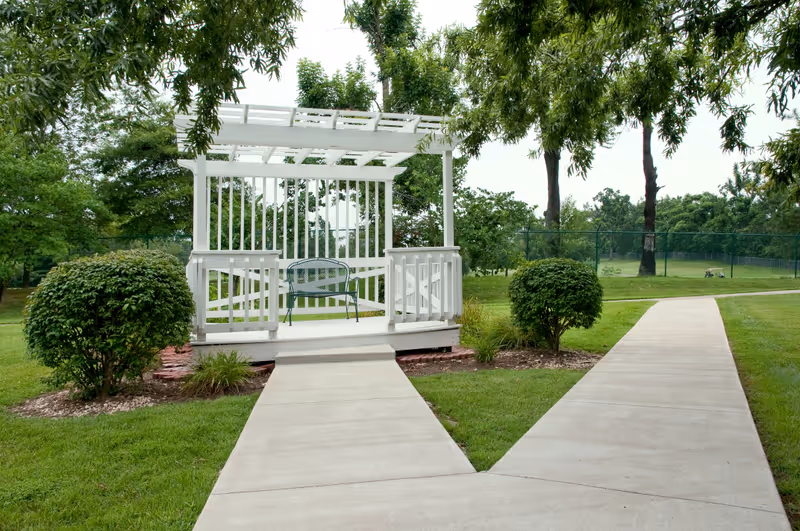 A white wooden gazebo with a bench inside, situated in a green outdoor area with trees, bushes, and a paved walkway leading to and past the gazebo.