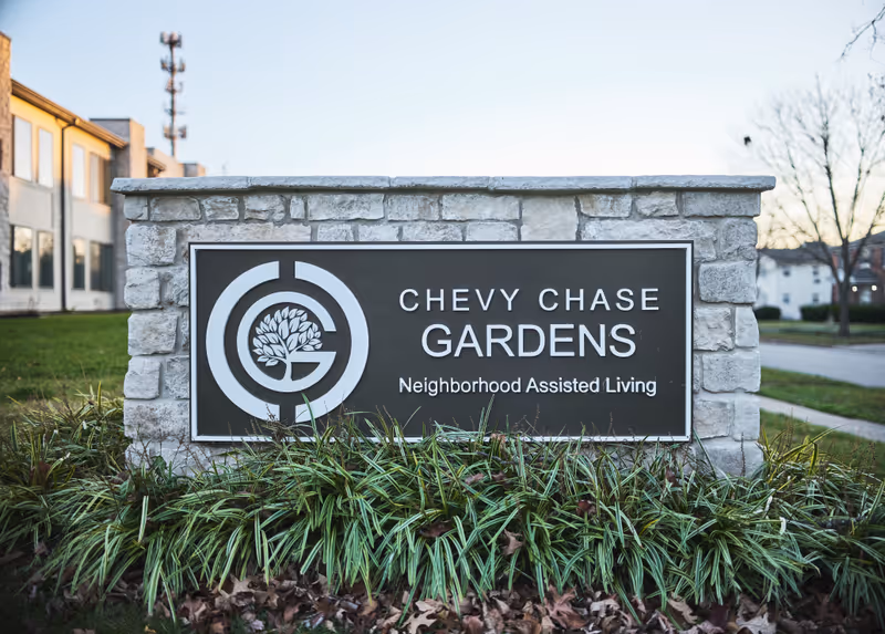 Stone entrance sign reads Chevy Chase Gardens, with landscaping, near a building facade.