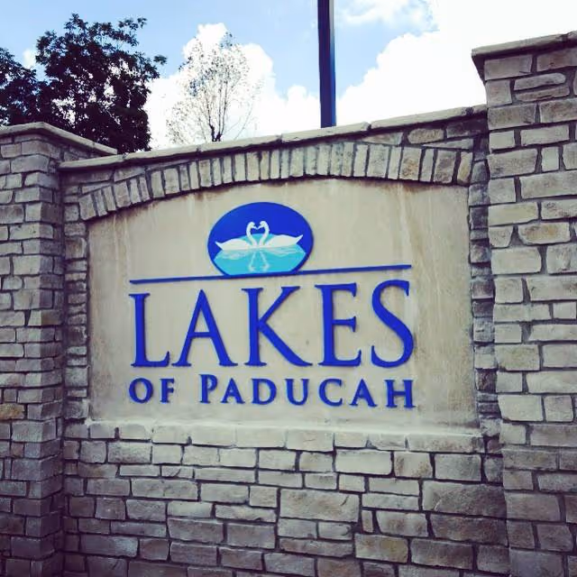 Stone entrance sign reading 'LAKES OF PADUCAH' with a swan logo set into a brick wall.