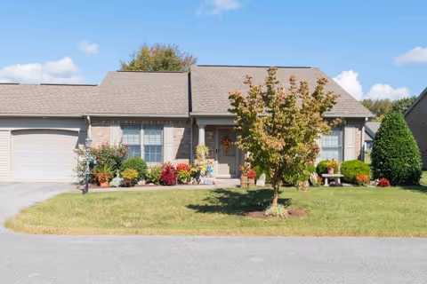 Single-story brick house with a gray roof, a small tree in the front yard, various plants and flowers near the entrance, and a driveway leading to a garage on the left side.