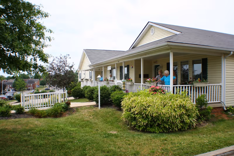 Single-story yellow assisted living building with a covered front porch, landscaping, and two people conversing on the porch.