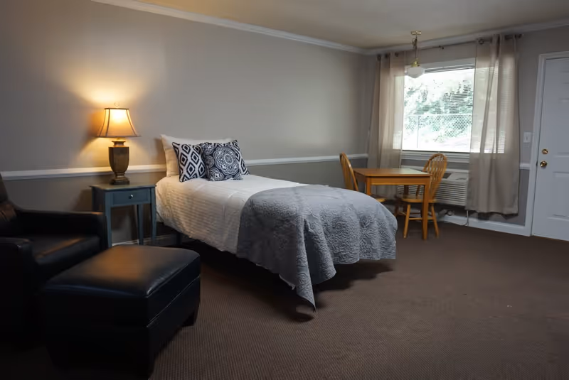 Furnished bedroom with a single bed, nightstand and lamp, armchair with ottoman, and a small table and chairs by a window.