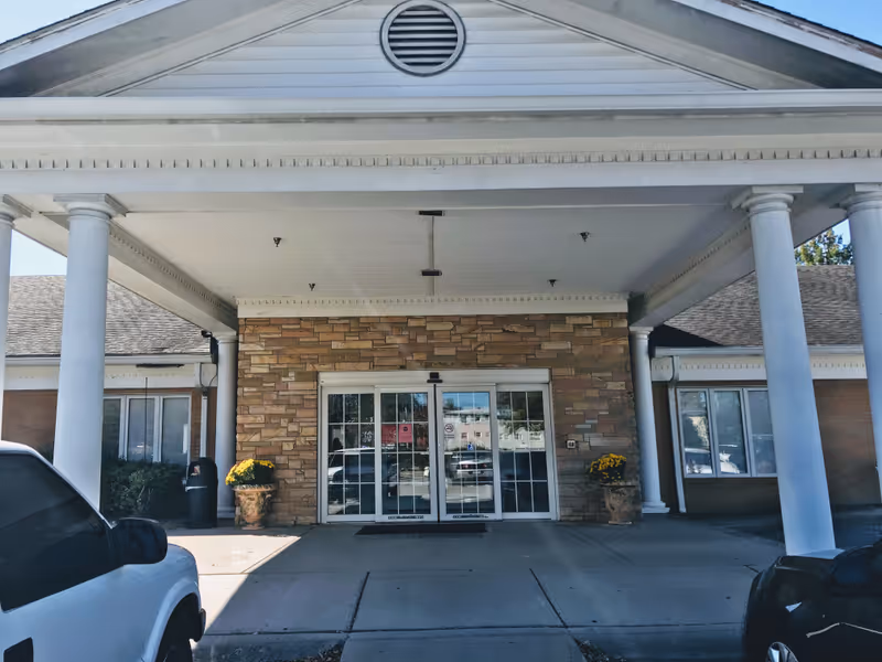 Front entrance of a senior living facility with a columned portico, stone facade, and glass double doors flanked by potted flowers.