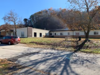 Single-story building with beige walls and a flat roof situated in a rural area with trees and hills in the background. A red car is parked on a paved area in front of the building, and there is a grassy area with a tree casting shadows in the foreground.
