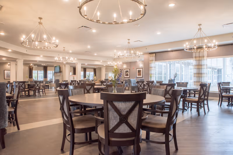 Spacious senior living dining room with multiple round and square tables surrounded by wooden chairs with cushioned seats. The room is well-lit with large circular chandeliers hanging from the ceiling and natural light coming through large windows with striped curtains. There are decorative flowers on the tables and framed artwork on the walls.