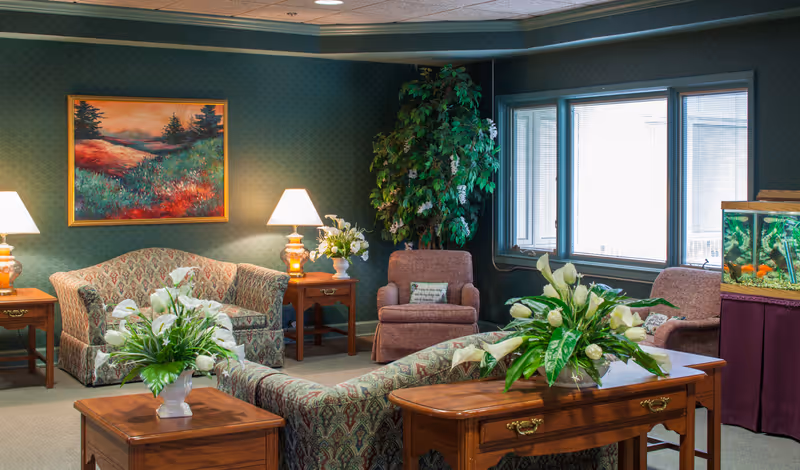 A cozy living room area with patterned sofas and armchairs, wooden side tables with lamps and flower arrangements, a large potted plant in the corner, a colorful landscape painting on the wall, and a fish tank near the window.