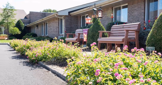 Flowerbed with pink blooms and wooden benches along the brick front of a senior living facility with windows and hanging bird feeders.