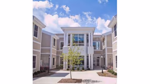 Two-story beige nursing home building with a central glass entrance and a small landscaped courtyard under a blue sky.