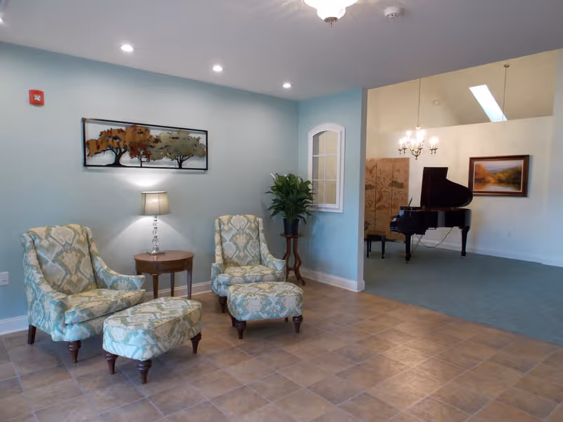 A seating area with two patterned chairs, ottoman, lamp, and piano in background.