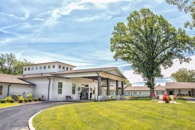 Exterior view of Dominion Senior Living At Northfield showing a single-story building with white brick walls and a covered entrance supported by wooden pillars. There is a large green lawn in front with a paved driveway curving towards the entrance. A large tree and some outdoor seating with red chairs and a flower pot are visible on the lawn under a partly cloudy sky.