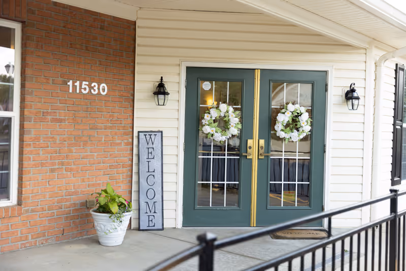 Double green glass entry doors with floral wreaths, a welcome sign, potted plant, and the street number 11530 on a brick exterior.