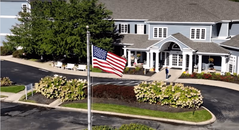 Exterior view of a senior living facility named Arcadia Louisville, showing a large building with white columns at the entrance, surrounded by landscaped bushes and trees. An American flag is prominently displayed on a flagpole in the foreground, and a person is walking near the entrance.