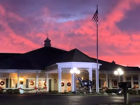 Exterior view of Crescent Place Assisted Living building at dusk with a vibrant pink and purple sky. The building is decorated with holiday wreaths and lights, featuring a covered entrance with white columns, a flagpole with an American flag, and a horse statue near the entrance.