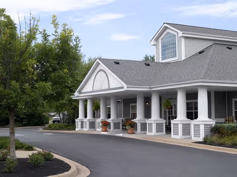 Covered entrance of a residential building with white columns, potted plants, and a driveway.