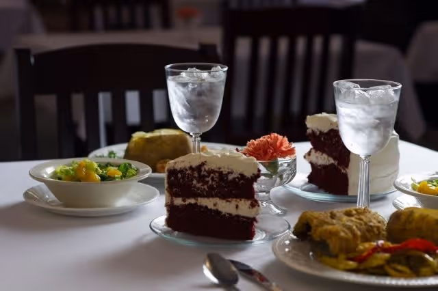 A dining table set with plates of food including salad, a main dish with vegetables, and slices of red velvet cake with white frosting. There are two glasses of water with ice and a small vase with a pink carnation flower on the table. The background shows dark wooden chairs.