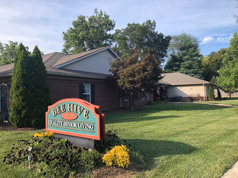 Front lawn and entrance of a Bee Hive senior living building with a brick facade and a prominent sign reading "Bee Hive Quality Senior Living".