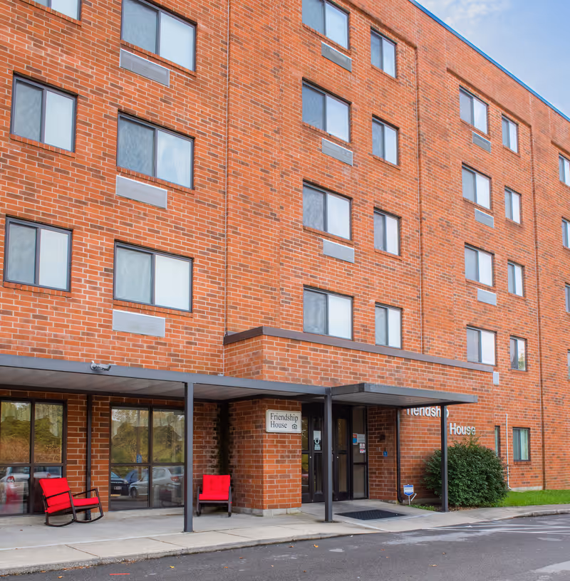 Exterior view of a multi-story brick building with several windows. The entrance has a covered walkway supported by metal poles. Two red cushioned chairs are placed near the entrance. A sign next to the door reads 'Friendship House'.