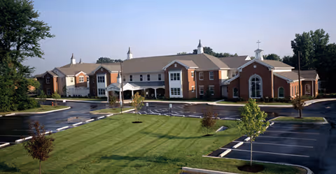 Exterior view of a large brick building with white trim and multiple peaked roofs, surrounded by a parking lot with marked spaces and small trees, under a clear blue sky.