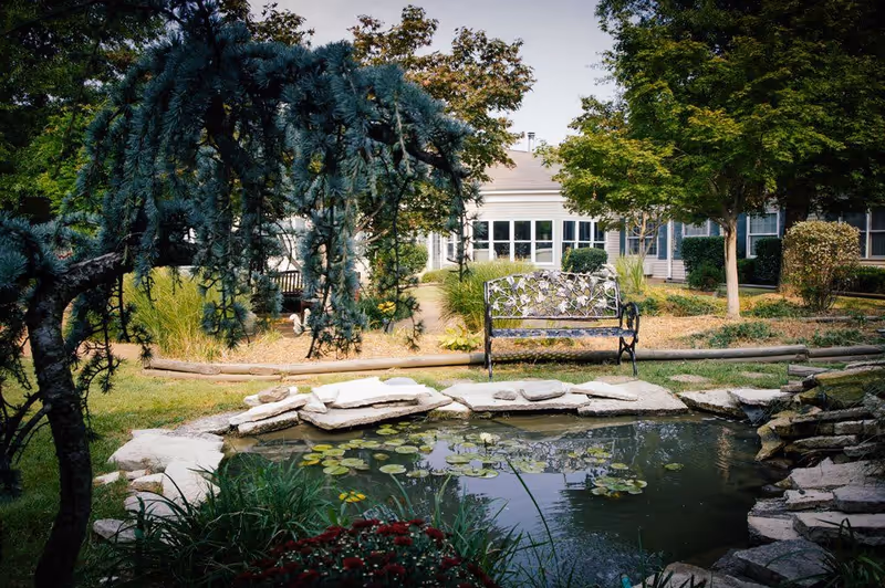 A peaceful outdoor garden area at Gaither Suites At West Park Assisted Living and Personal Care Home featuring a small pond with lily pads surrounded by rocks, a decorative metal bench, trees, shrubs, and a building with large windows in the background.
