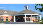 Front entrance of a senior living building with a covered porte-cochère, white columns, and brick facade under a blue sky.