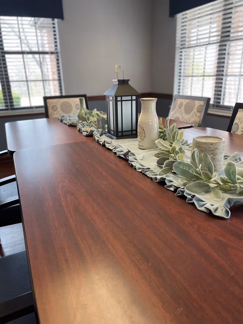Long wooden dining table with a lantern centerpiece and green foliage in a bright dining room.