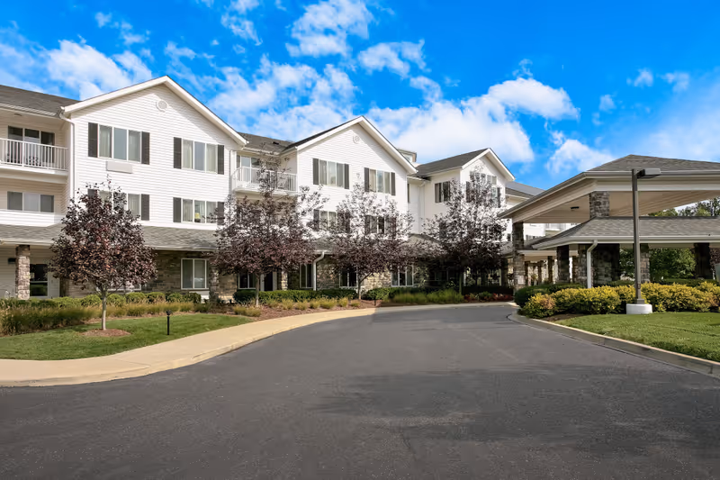 Exterior view of a multi-story senior living facility building with white siding and stone accents. The building has multiple windows and balconies, with a covered entrance driveway surrounded by landscaped bushes and small trees under a partly cloudy blue sky.