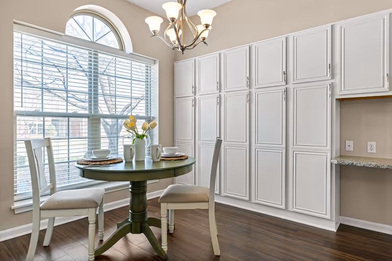 A bright dining area with a round green table set for two with white chairs. The table has a vase with yellow and white tulips, two white mugs, and two bowls on woven placemats. Behind the table is a large window with white blinds and an arched top, letting in natural light. To the right, there is a wall with multiple white cabinets and a small granite countertop with electrical outlets above it. The floor is dark wood, and a chandelier with five lights hangs above the table.