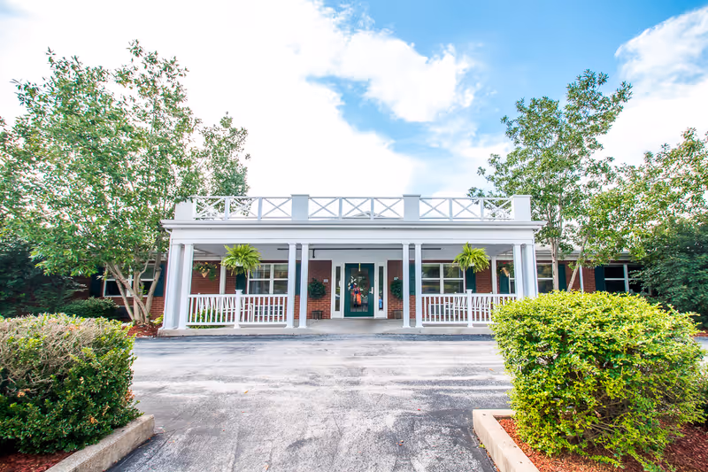 Front entrance of a single-story brick care facility with a white columned porch, green door, hanging plants and surrounding trees.