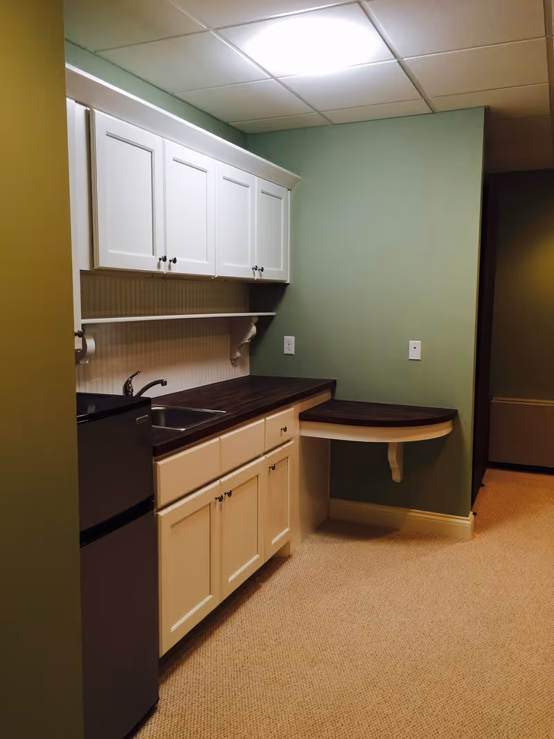 Small kitchenette with white cabinets, a dark wood countertop, sink, mini fridge, and a fold-down table against green walls.