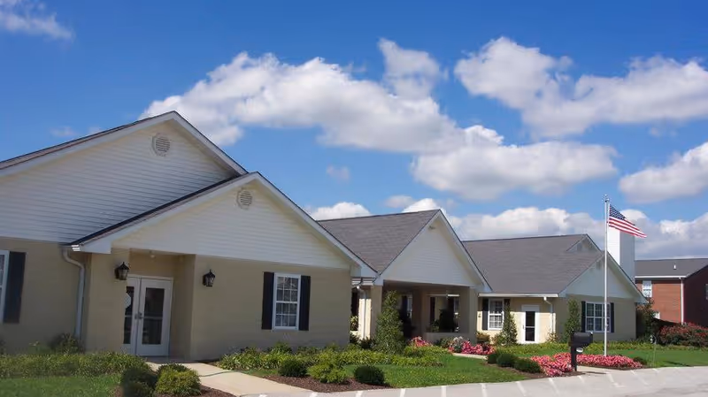 Front exterior of a single-story assisted living building with gabled roofs, landscaped grounds, and an American flag.