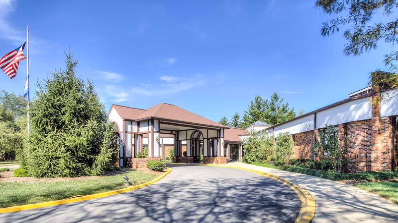 Exterior view of The Forum at Brookside senior living facility with a driveway leading to the entrance. The building has a combination of brick and white paneling with brown trim. There are trees and shrubs around the building, and an American flag is visible on a flagpole to the left. The sky is clear and blue.