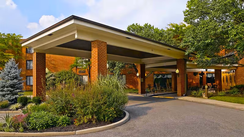 Entrance of a senior living facility with a covered drop-off area supported by brick pillars, surrounded by landscaped greenery including bushes and trees, with a brick building in the background under a partly cloudy sky.