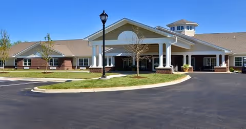 Front exterior of a single-story senior living facility with a covered entrance, circular driveway, lamp post, and clear blue sky.
